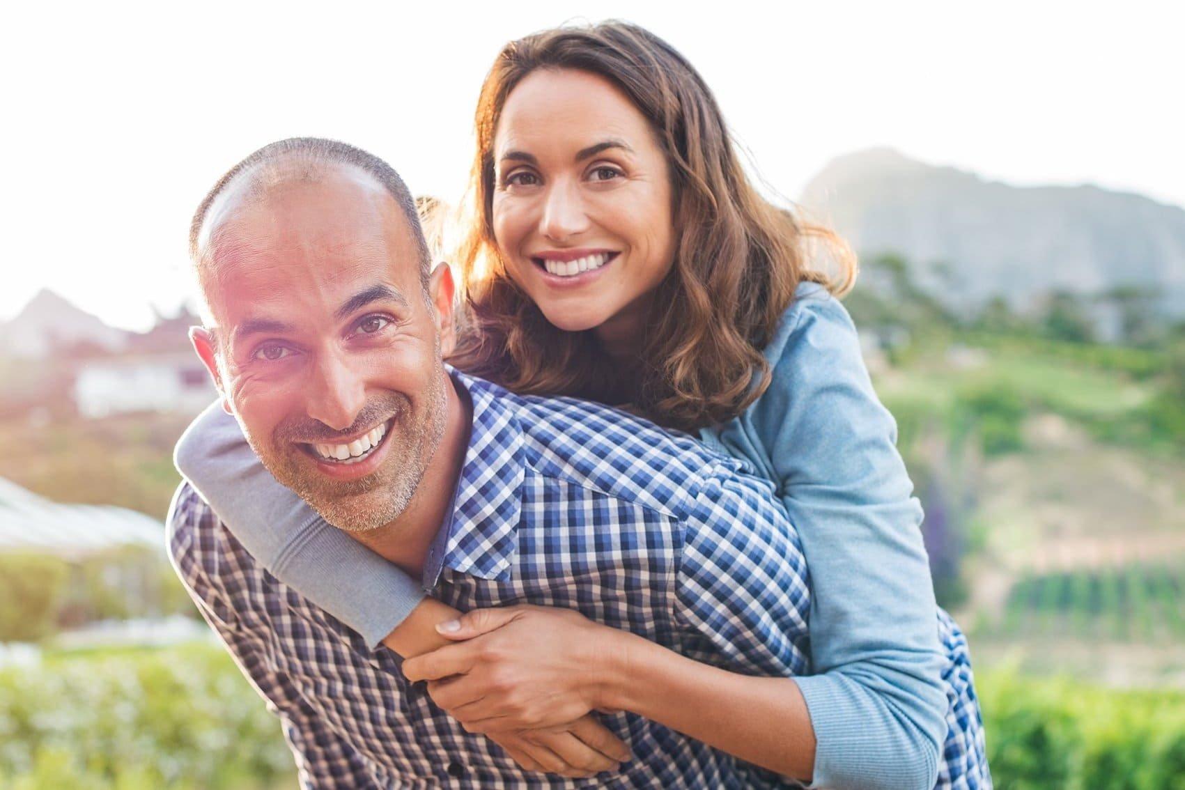 A smiling woman gives a piggyback ride to a smiling man outdoors, with greenery and mountains blurred in the background. Both look happy and relaxed, enjoying a bright, sunny day—perhaps thanks to ICL for those with high prescriptions.