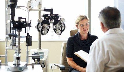 A woman sits in an eye exam room, attentively listening to a male optometrist discuss LASIK options. Ophthalmic equipment and a phoropter are visible beside them. Natural light enters through a window in the background.