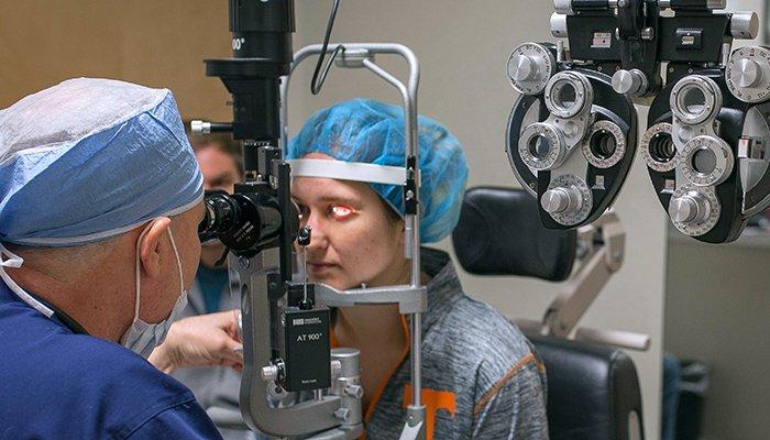 A patient wearing a blue hair cover undergoes an eye exam for LASIK, with an ophthalmologist using a slit lamp while eye examination equipment is visible in the foreground.