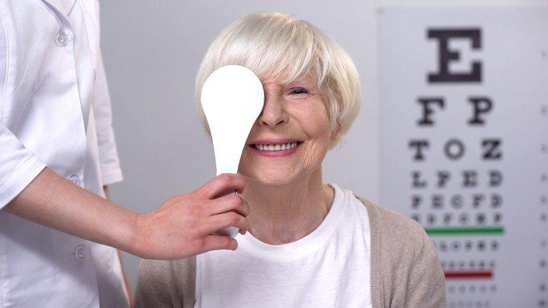 Smiling elderly woman at an eye exam, covering one eye with a paddle as a healthcare professional assists her; an eye chart is visible in the background, highlighting the importance of regular checkups before considering laser eye surgery.