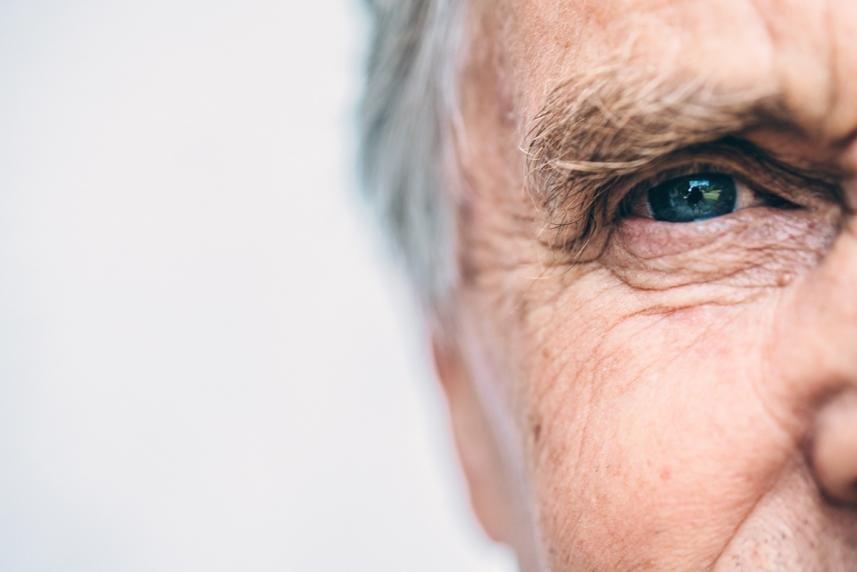Close-up of an older man's face, focusing on his blue eye and the wrinkles around it—capturing detail reminiscent of topography-guided laser correction—while the left side fades out of frame against a light, blurred background.