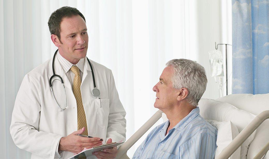 A doctor with a stethoscope and clipboard talks to an older man in a hospital bed, discussing laser eye surgery. The man, wearing blue-striped pajamas, listens attentively to the doctor's explanation.