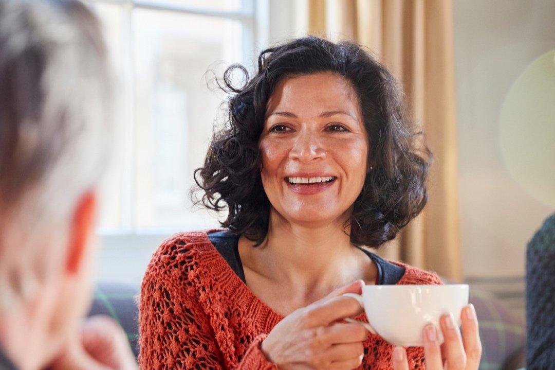 A woman with curly dark hair wearing an orange sweater smiles warmly while holding a white mug, sitting indoors near a window. She discusses implantable contact lenses for high prescriptions. Another person is partially visible in the foreground.
