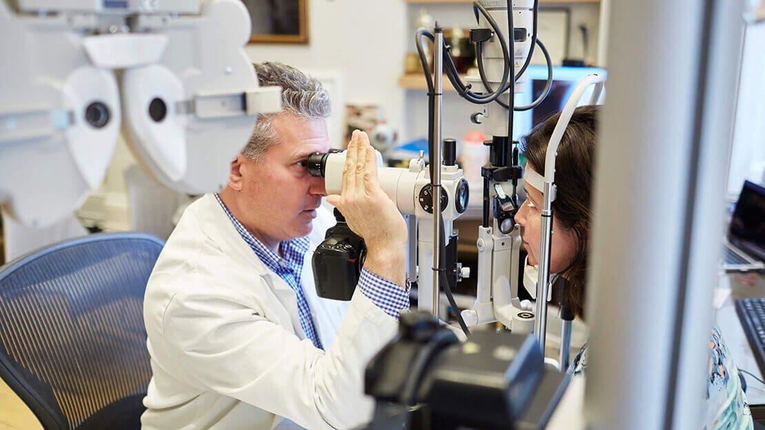 An eye doctor in a white coat examines a patient's eyes using a slit lamp microscope in a medical office, discussing LASIK and PRK as possible vision correction options. The patient is seated with her chin on the device's rest.