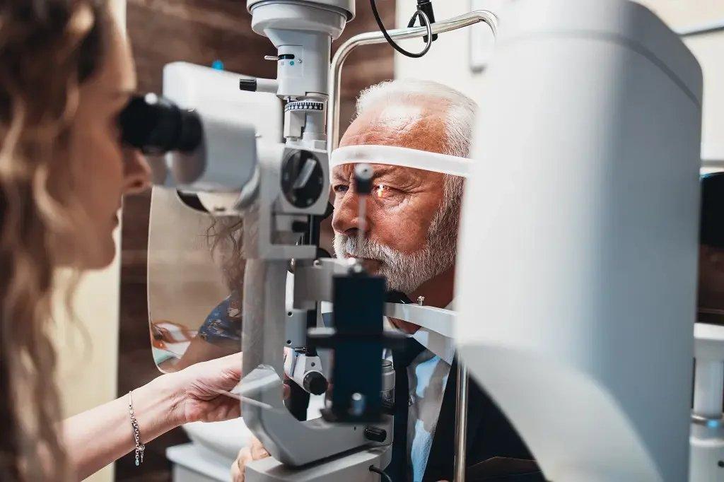 An older man with gray hair and beard undergoes an eye examination, possibly as a pre-screening for laser eye surgery, looking into a medical device operated by a healthcare professional in a clinical setting.