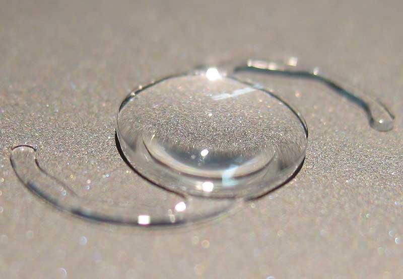 A close-up of a transparent Rayner Galaxy IOL placed on a glittery surface, showing its curved center and two thin, curved arms extending from each side—designed to deliver high-quality vision.