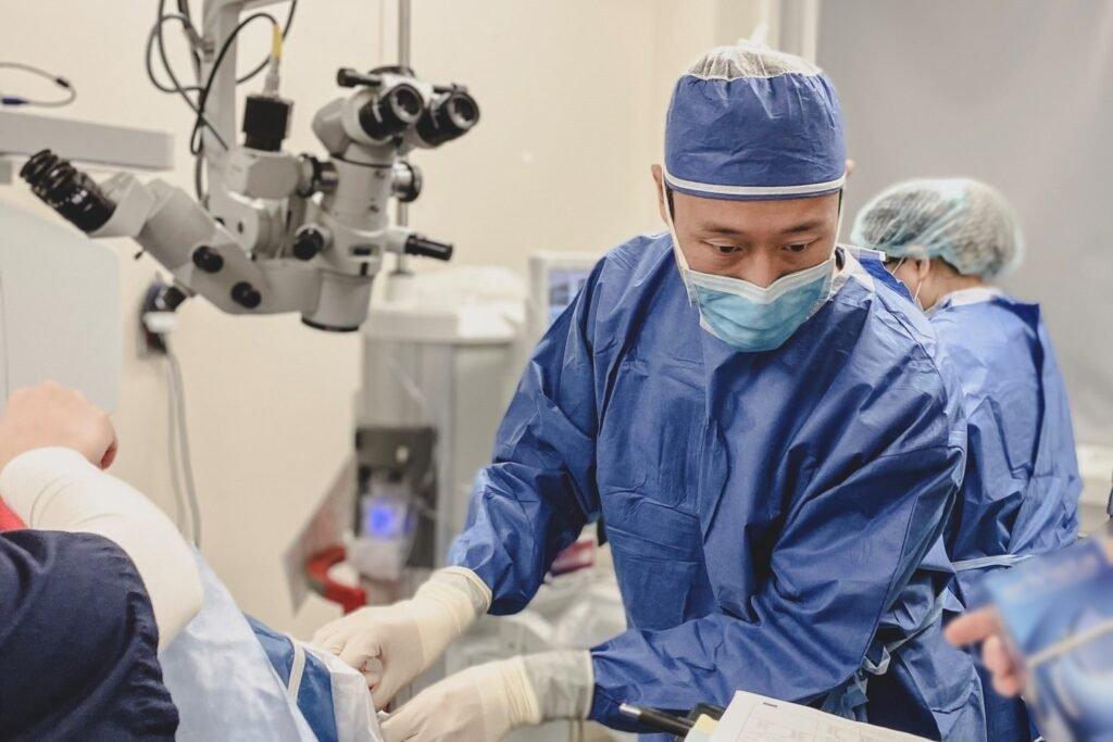 A surgeon in blue scrubs and a mask performs implantable contact lens surgery in an operating room, with surgical equipment and another medical professional visible in the background.