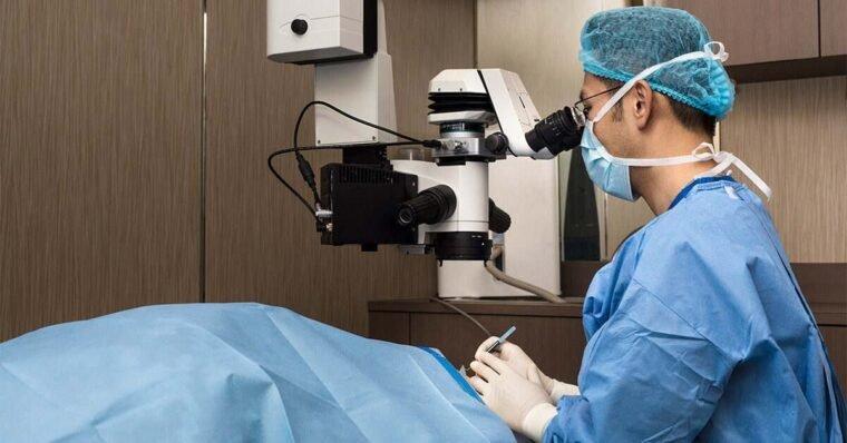 A surgeon in blue scrubs, a hair cover, and a face mask performs LASIK eye surgery using a microscope on a patient covered with a blue drape in a medical facility.
