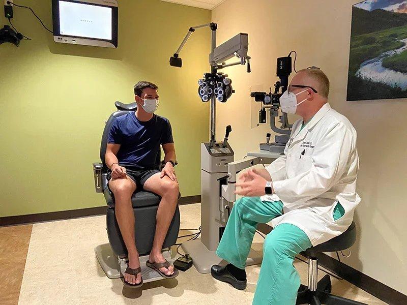 A patient sits in an examination chair while an eye doctor in a white coat and mask discusses SMILE or LASIK options with him in an eye clinic room equipped with vision testing devices.