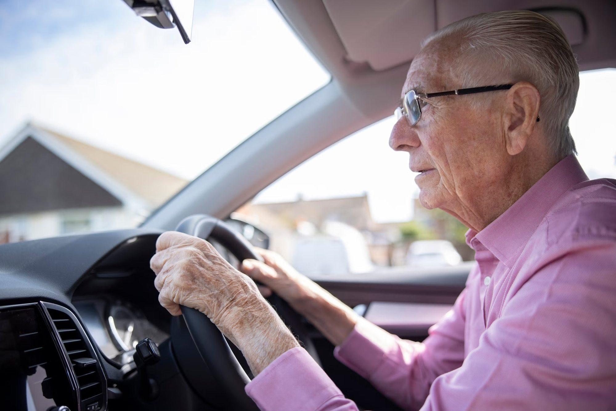 An elderly man wearing glasses and a pink shirt is driving a car, looking attentively out the windshield with both hands on the steering wheel, demonstrating careful driving after pterygium surgery. Sunlight streams through the window.
