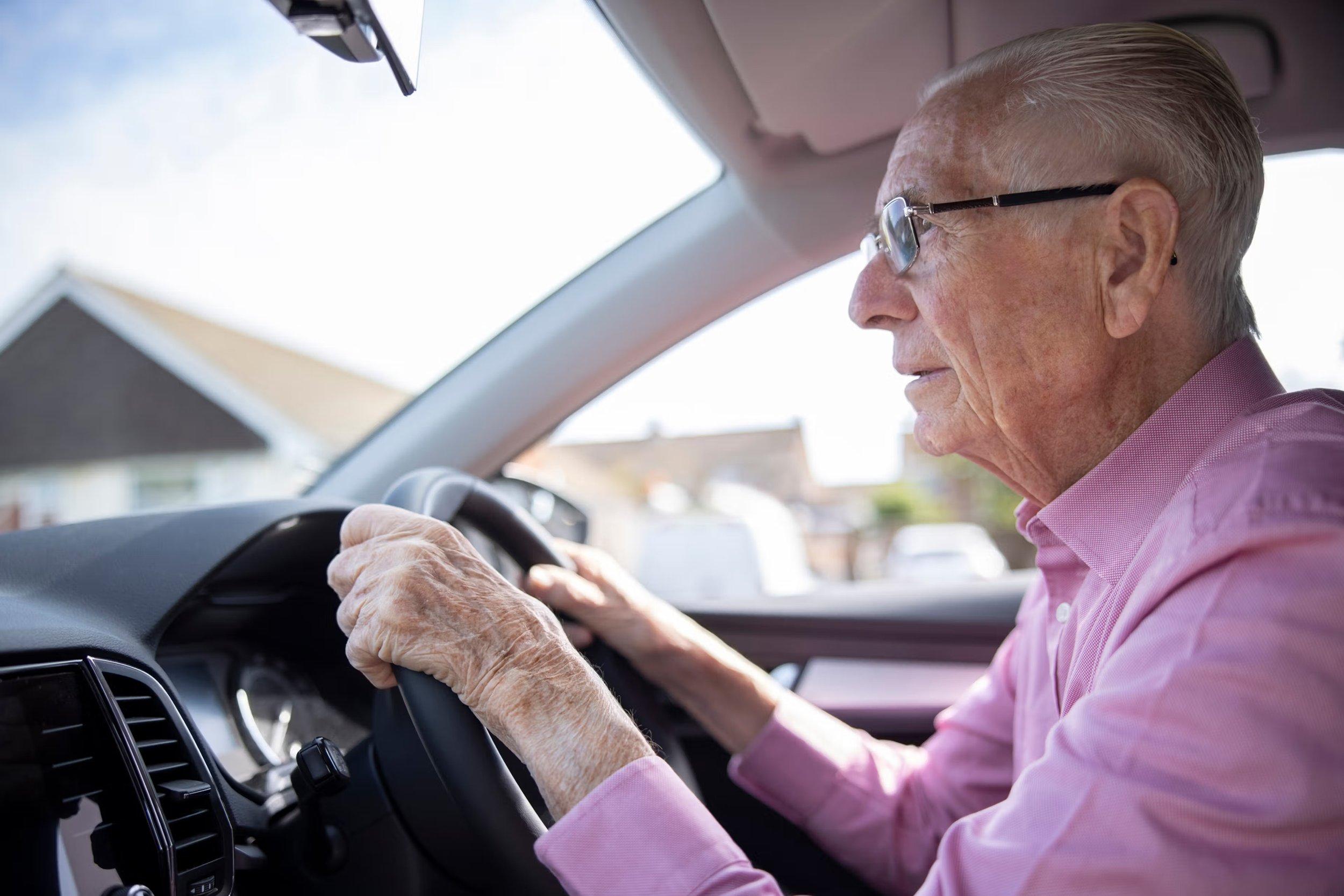 An elderly man wearing glasses and a pink shirt is driving a car, looking attentively out the windshield with both hands on the steering wheel, demonstrating careful driving after pterygium surgery. Sunlight streams through the window.