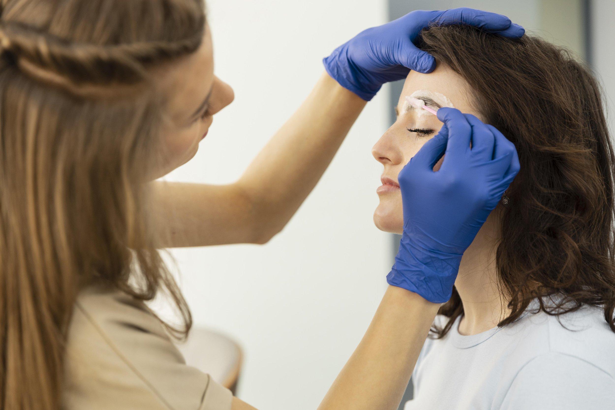 A beauty technician wearing blue gloves carefully shapes a woman’s eyebrow using tweezers, while discussing lens flexibility for presbyopia treatment as the woman sits with her eyes closed.