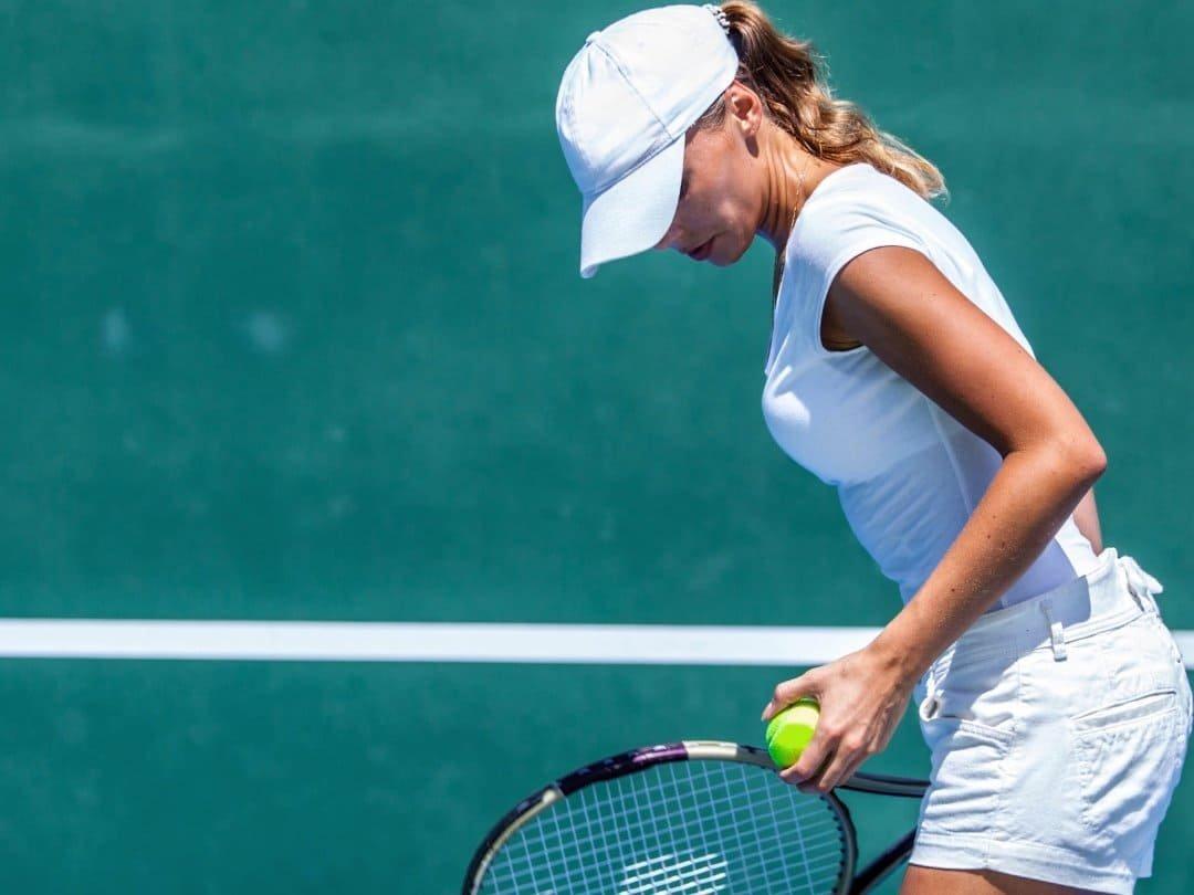 A woman in a white outfit and cap stands on a tennis court, looking down and holding a tennis ball and racket, enjoying clear vision as she prepares for her next move against the green background.