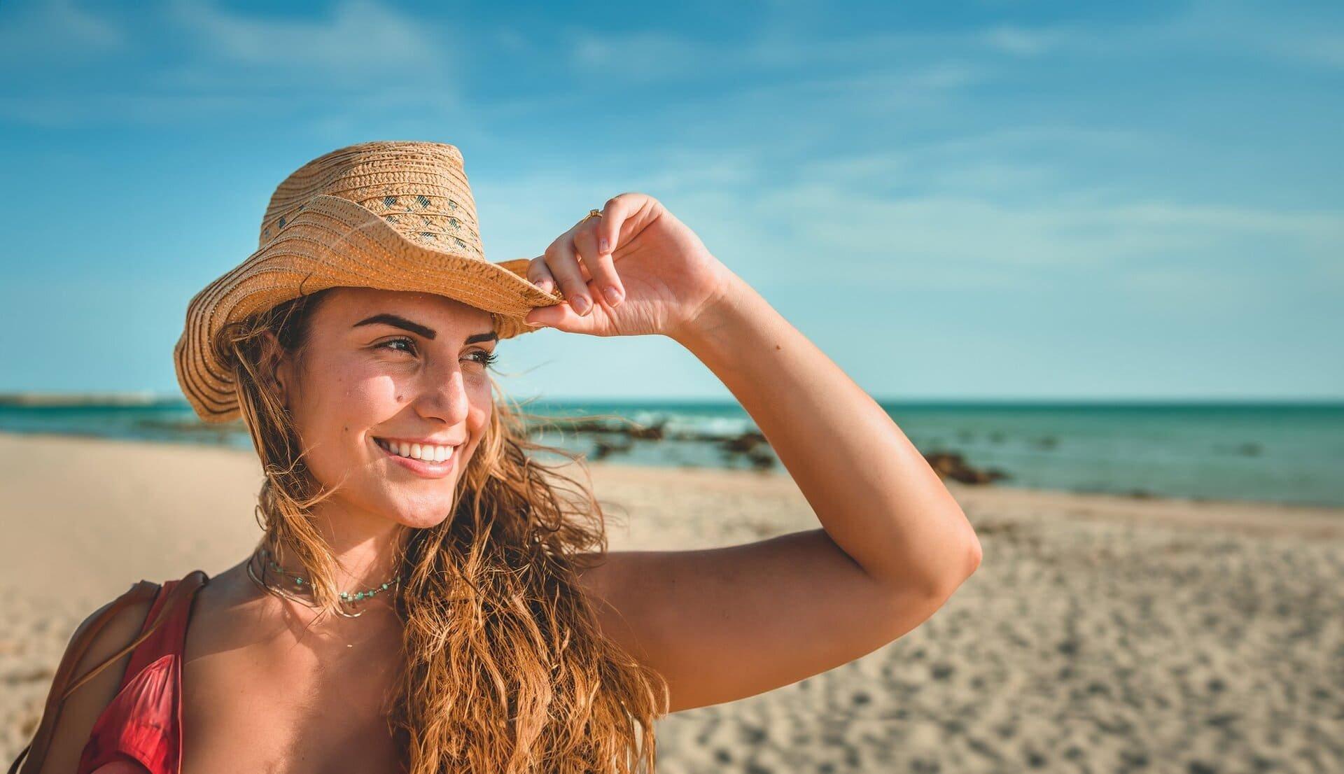 A smiling woman in a straw hat on a sunny beach, enjoying clear vision without glasses.