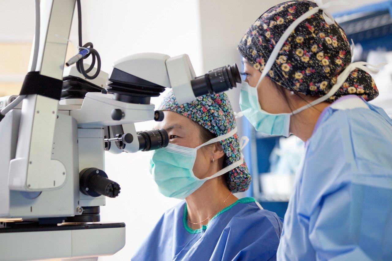 Two medical professionals in surgical scrubs, face masks, and floral-patterned caps use a microscope in a clinical setting, closely examining details as they perform an implantable contact lens surgery together.
