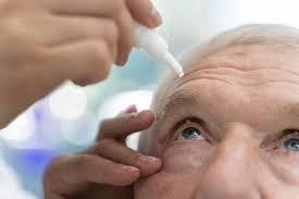 A close-up of an elderly person having eye drops administered, with one hand gently holding their eyelid open and the other holding the dropper above their eye, a common step before cataract surgery.