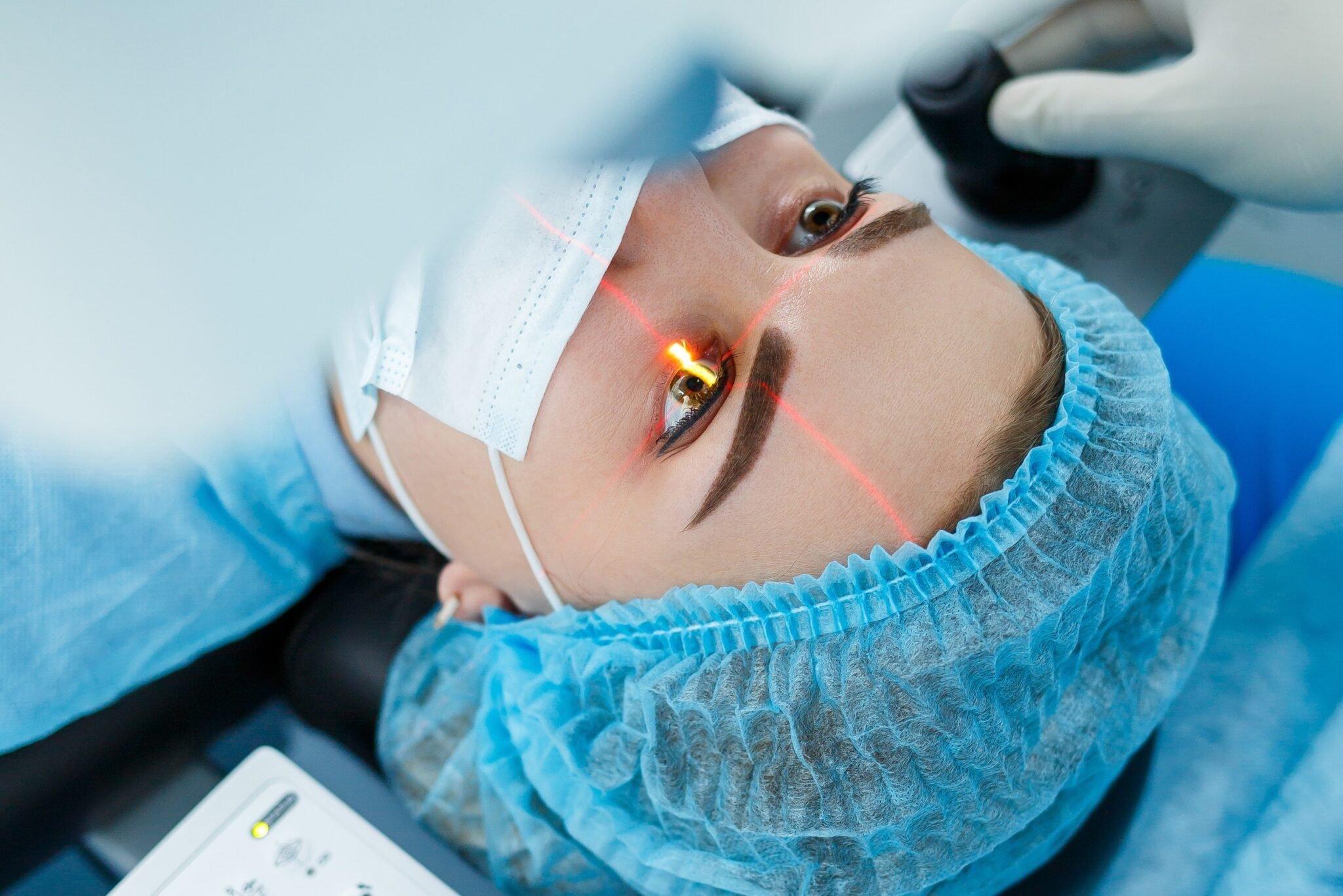 A person wearing a surgical mask and cap undergoes laser eye surgery, lying down as a red laser beam is precisely directed at their eye by a gloved medical professional, aiming to achieve crystal-clear vision.