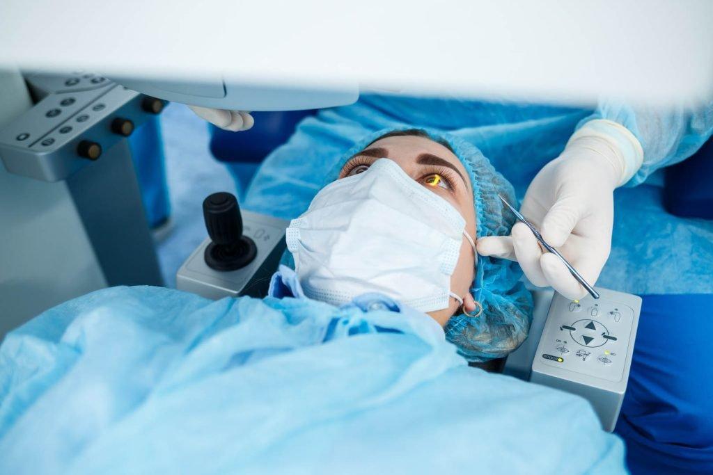 A patient wearing a surgical cap and mask lies on an operating table during LASIK eye surgery, with a medical professional using tools near the patient's eye for vision correction.