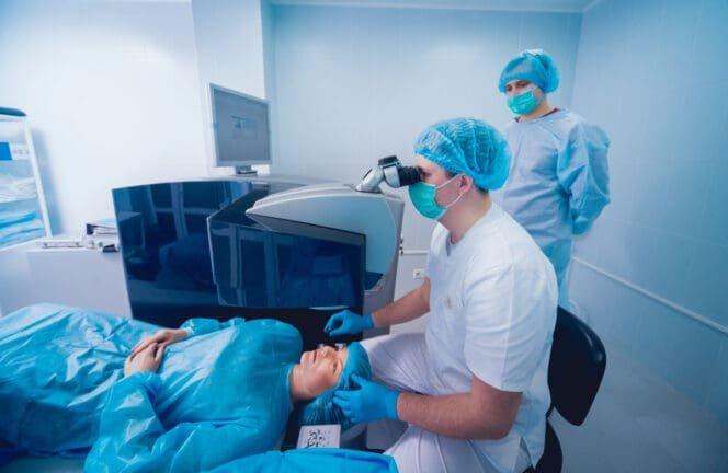 A patient lies on an operating table while a doctor in scrubs, gloves, and a mask performs cataract eye surgery using specialized equipment. Another medical professional observes in the background, illustrating how long cataract surgery takes.