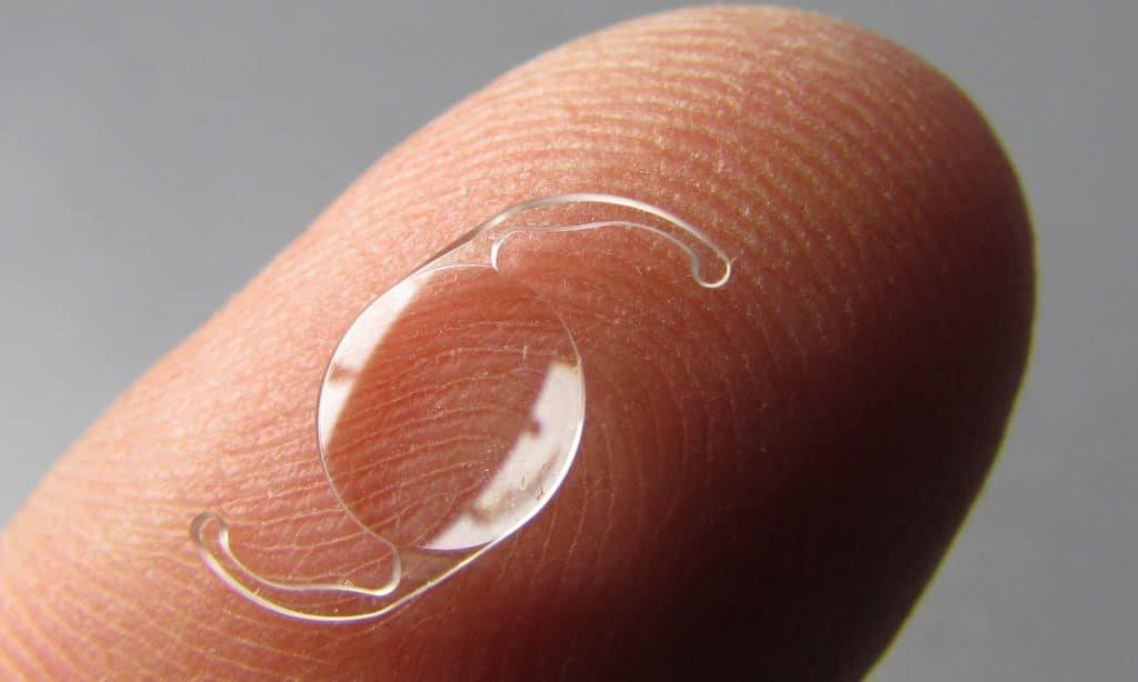 A close-up of a transparent Rayner Galaxy IOL resting on the tip of a finger, showing its small, curved shape against the skin’s detailed fingerprint for high-quality vision.
