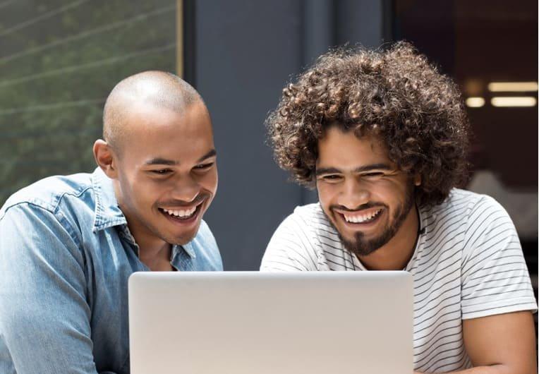 Two men sitting side by side, smiling and looking at a laptop screen as they research the benefits of laser eye surgery.