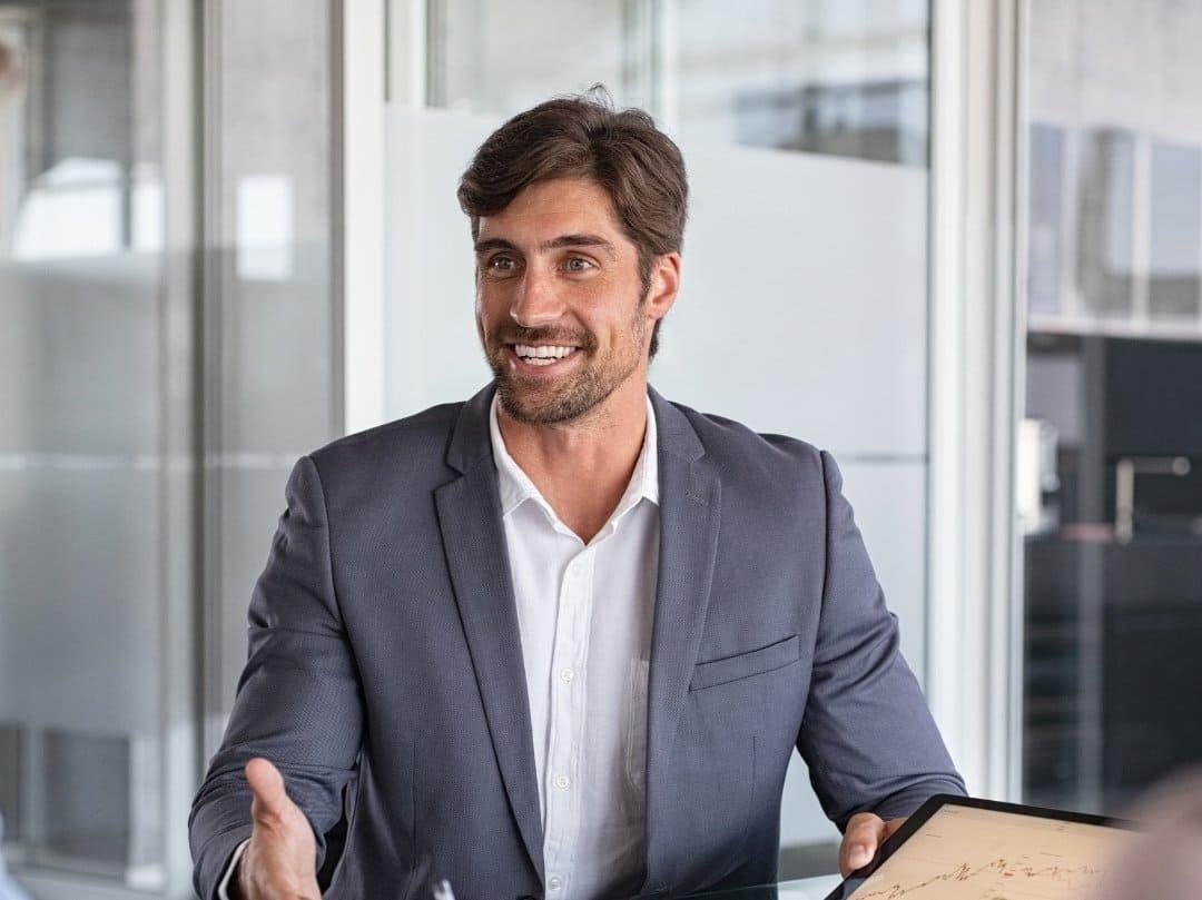 A man in a suit smiles while sitting at a desk in a modern office, gesturing with one hand and holding a tablet, ready to discuss your free assessment for LASIK surgery.