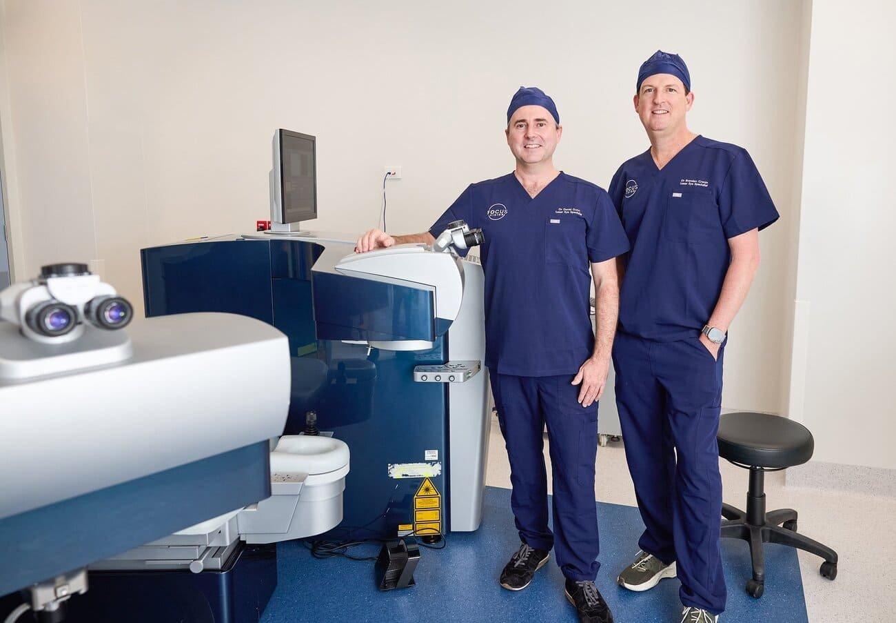 Two male medical professionals in blue scrubs stand in a modern clinic beside advanced ophthalmology equipment, ready to perform laser cataract surgery and offer tailored IOL solutions for clearer vision.