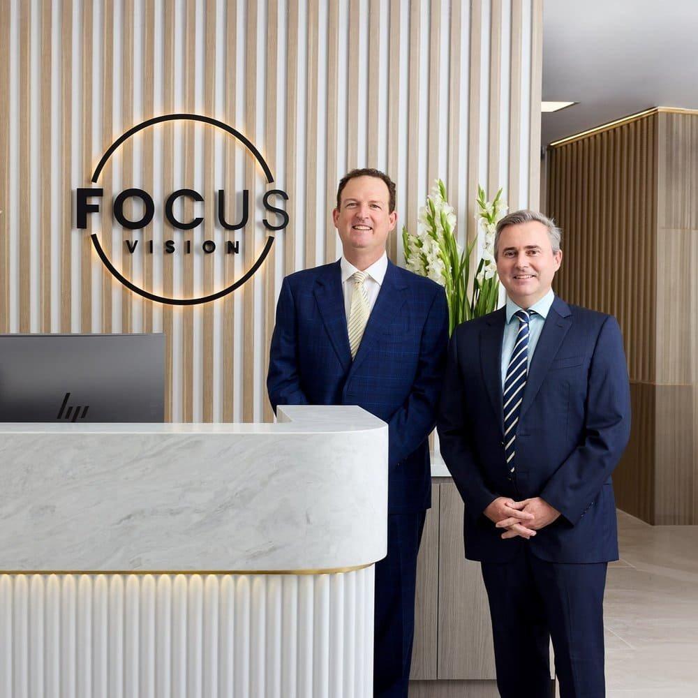 Two men in suits stand smiling at a modern reception desk with a Focus Vision sign on a striped wall behind them, representing leading ophthalmologists in laser eye surgery Brisbane. Tall white flowers decorate the corner of the counter.
