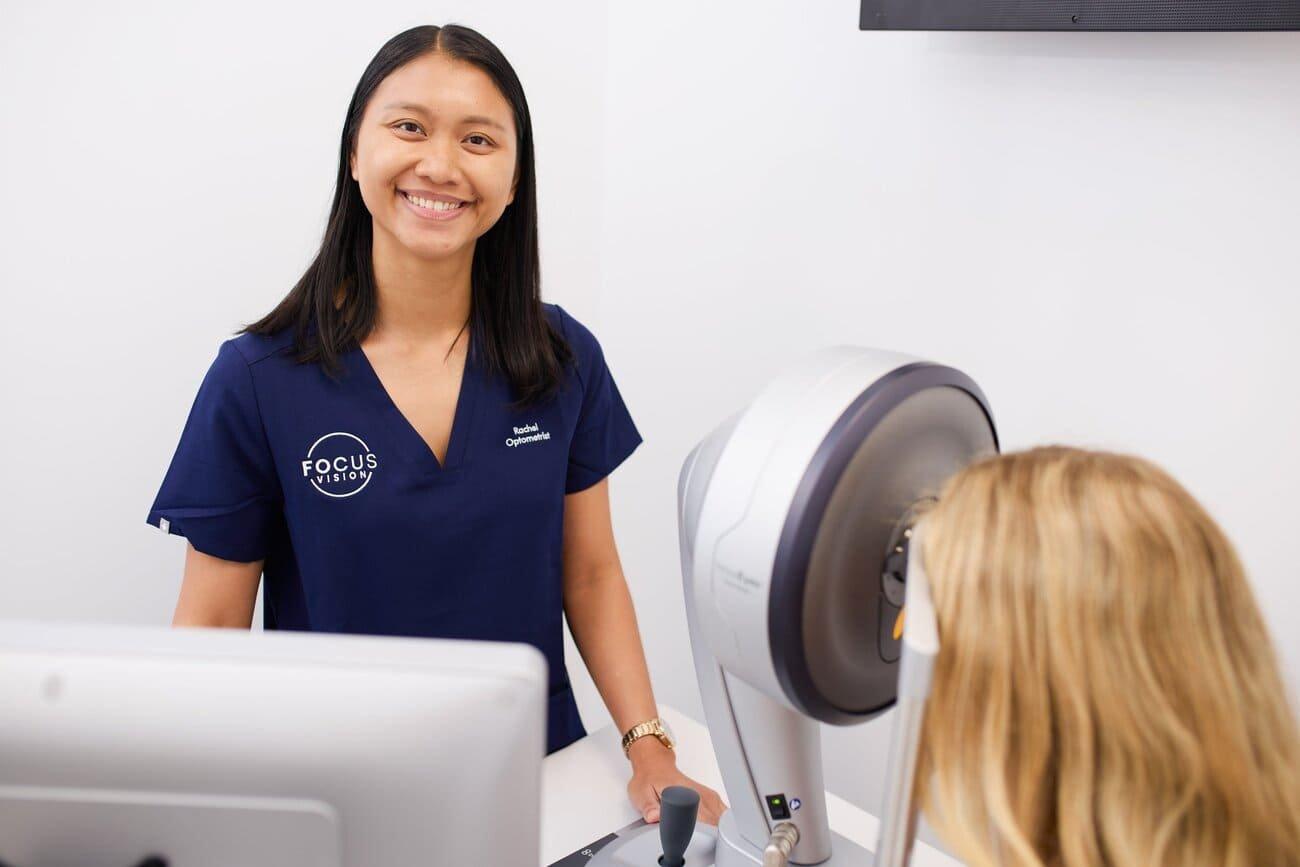 A smiling optometrist in navy scrubs stands beside eye exam equipment while a patient with blonde hair undergoes an eye test in a clean, white clinic—perfect for those considering laser eye surgery with a stable prescription.