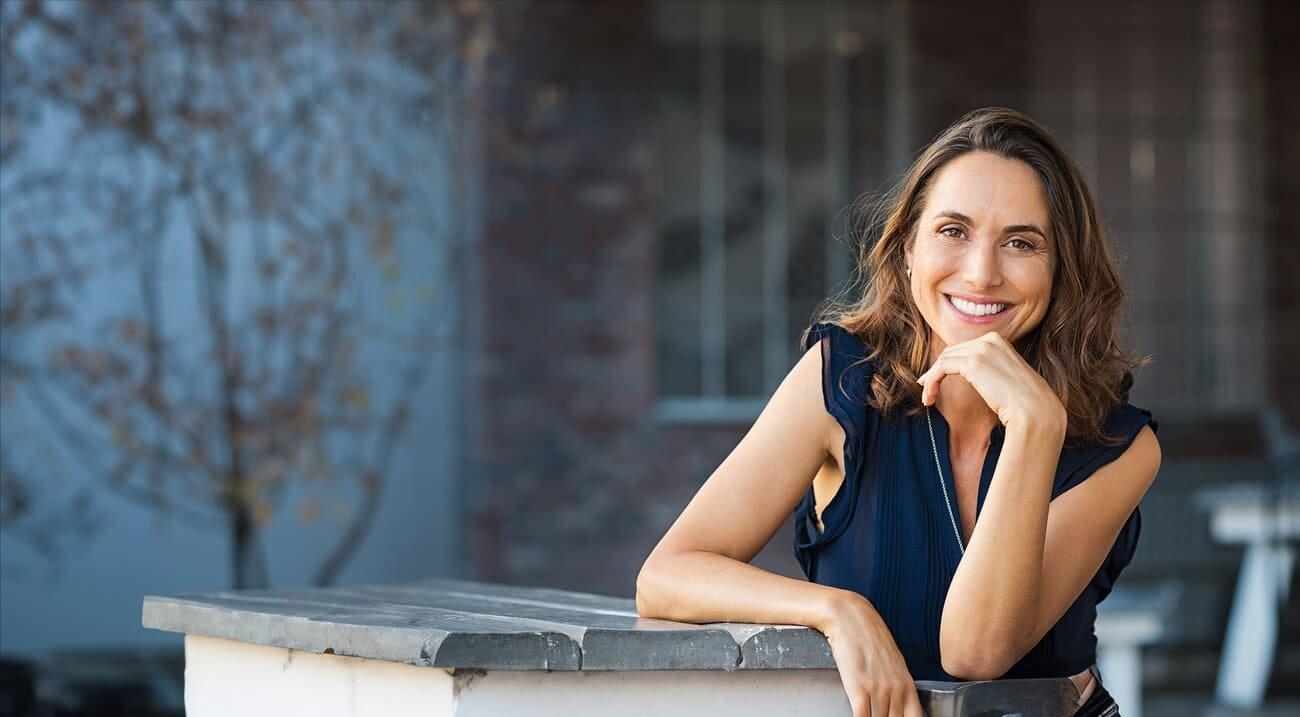 A woman with long brown hair wearing a navy blouse sits outdoors at a stone table, smiling and resting her chin on her hand. Considering laser eye surgery? Book your consult if you have a stable prescription. Trees and a brick wall blur in the background.