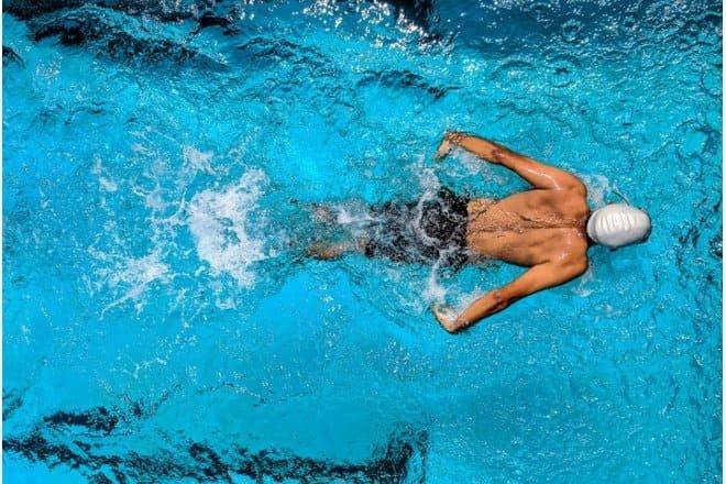 A swimmer wearing a white swim cap and goggles, who recently underwent an ICL procedure, swims freestyle in a bright blue pool, creating splashes as they move forward, viewed from above.