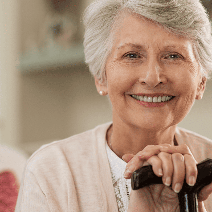 Smiling elderly woman with short gray hair, wearing a light cardigan, rests her hands on a black walking cane and looks at the camera in a cozy indoor setting, enjoying clearer vision after laser cataract surgery.