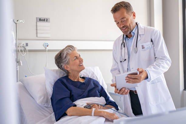 A doctor stands beside a hospital bed, smiling and talking to a female patient in a hospital gown. The patient is smiling back as the doctor holds a tablet, explaining how long cataract eye surgery takes and what to expect during recovery.