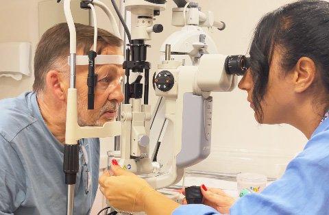 A doctor in a blue coat examines an older man's eyes using a slit lamp biomicroscope during an eye exam, discussing possible LASIK or PRK procedures in a medical office.