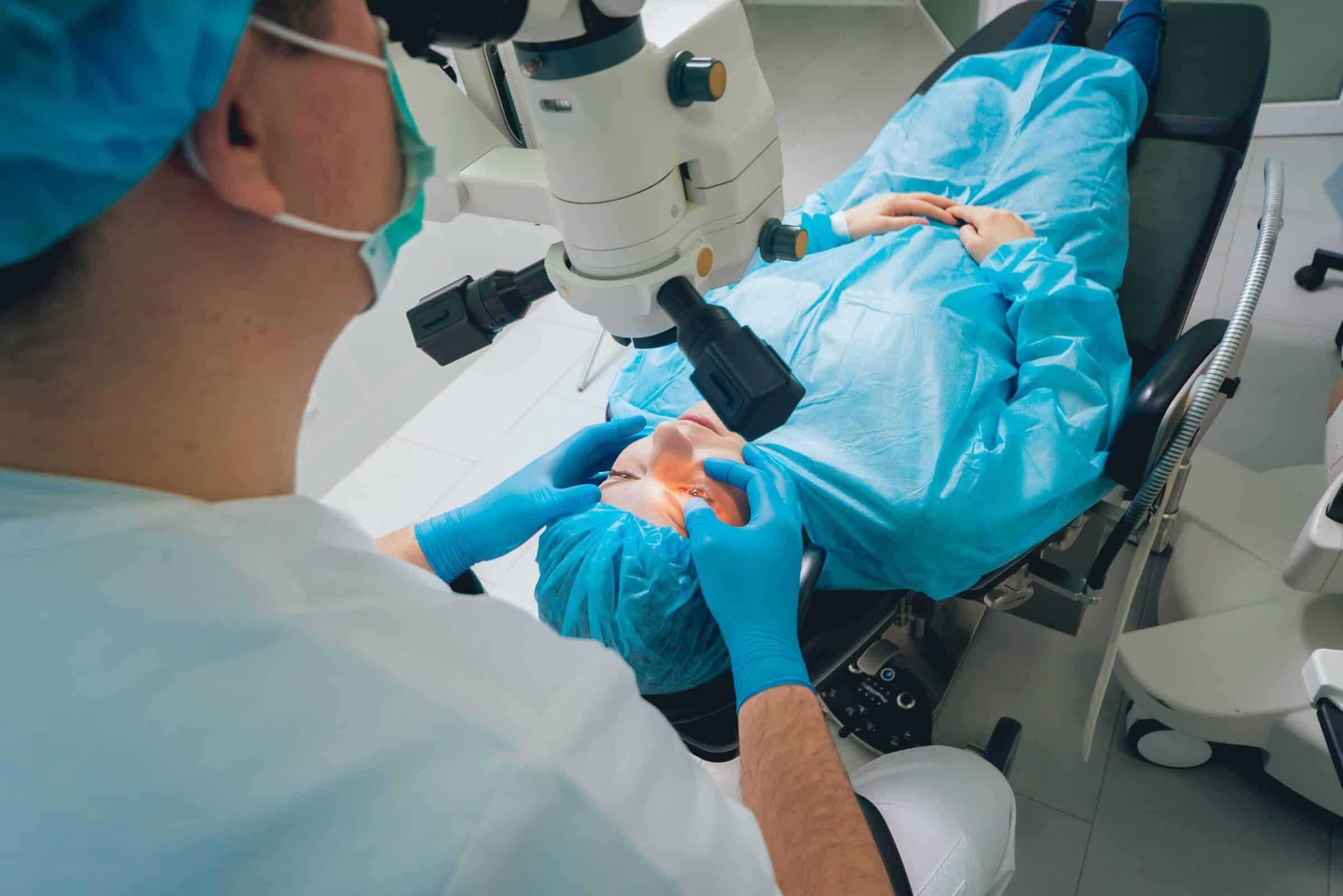 A patient lies on a medical chair covered in blue surgical drapes while a doctor in scrubs and gloves performs LASIK eye surgery using a large microscope.