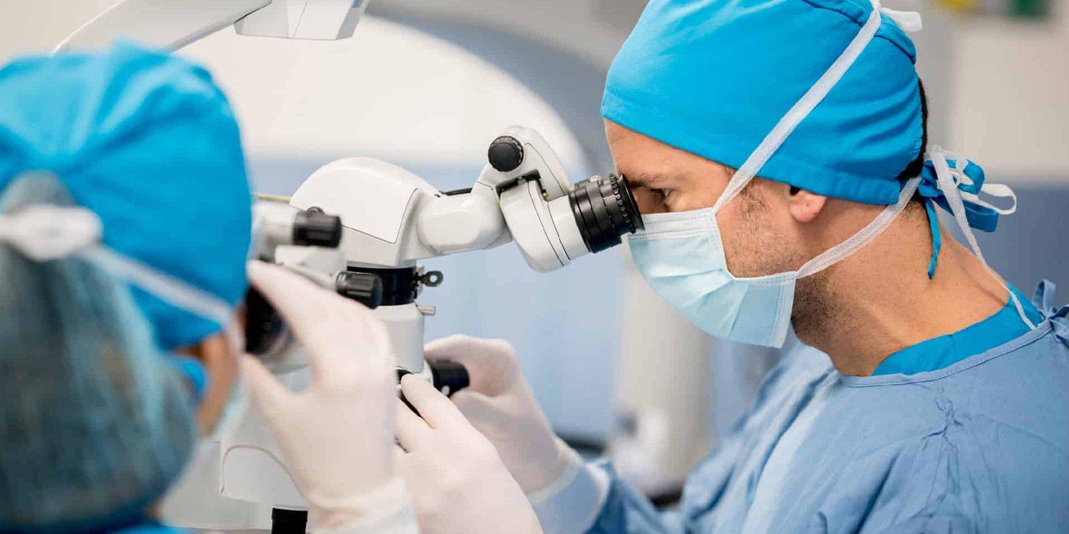 A surgeon in blue scrubs, gloves, mask, and cap uses a microscope during LASIK eye surgery. Another medical professional, also in scrubs, is visible in the foreground assisting with the laser eye surgery procedure.