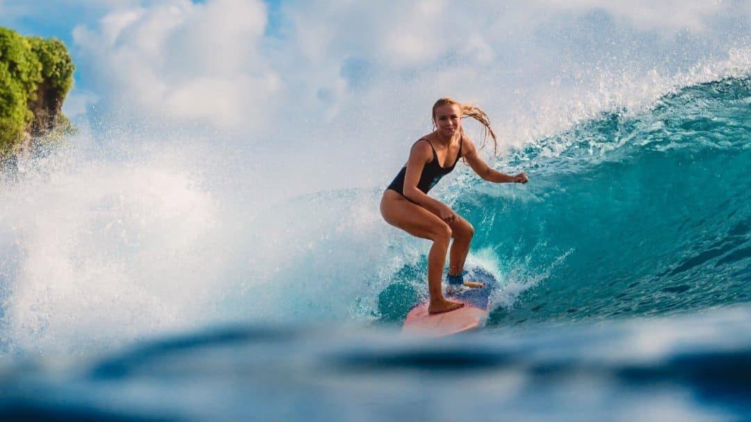 A woman in a black swimsuit rides a turquoise wave on a surfboard, with a rocky green cliff behind her.