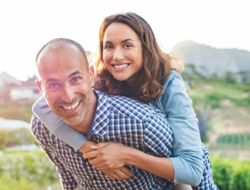 A smiling man gives a piggyback ride to a woman outdoors. They both look happy, enjoying the greenery and hills in the background—perhaps celebrating the joy of clearer vision after LASIK surgery.
