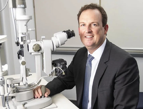 Dr Brendan Cronin in a suit sits beside an ophthalmology slit lamp microscope in a medical office, smiling at the camera—symbolizing expertise in advanced LASIK and commitment to better vision.