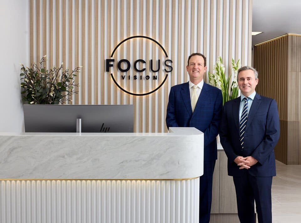 Dr Brendan Cronin and Dr David Gunn in suits standing and smiling behind a marble reception desk with a computer, in front of the FOCUS VISION logo and modern wood paneling. Green plants decorate the area, reflecting their commitment to quality eye care Brisbane.