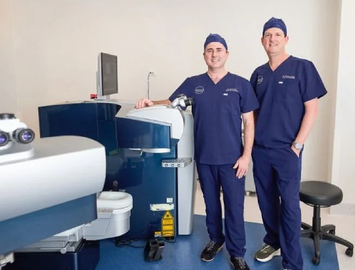Two male medical professionals (Dr Brendan Cronin & Dr David Gunn) in blue scrubs and caps stand next to advanced LASIK equipment in a clean, modern clinic room. A stool and computer monitor are also visible in the background, highlighting a setting dedicated to clearer vision.