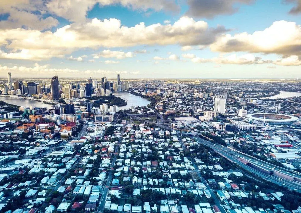 Aerial view of Brisbane cityscape with skyscrapers, river, suburban housing, and a sports stadium under a partly cloudy sky.